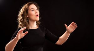 A woman with brown hair and a black shirt singing with upraised hands